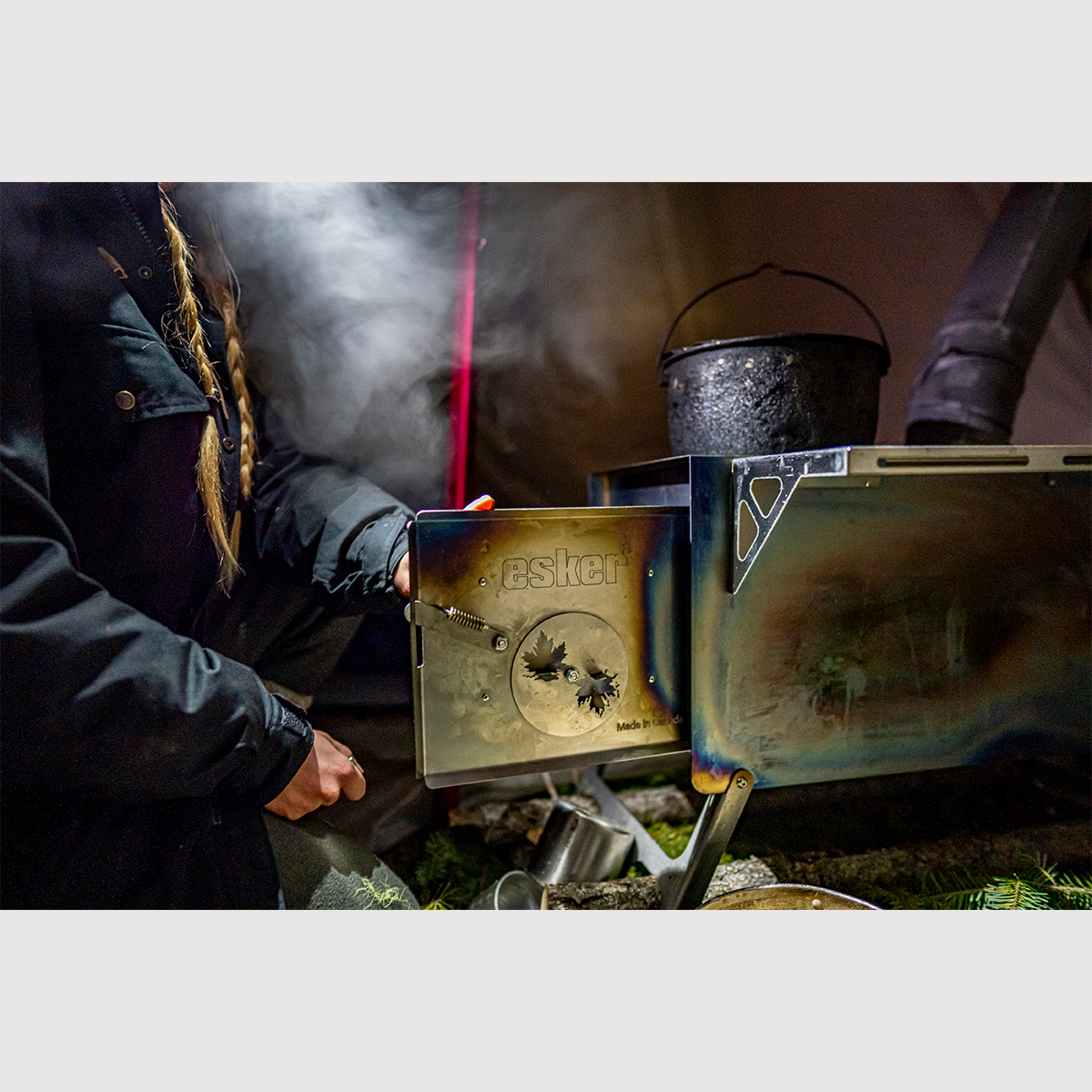 Person using an Esker portable Wood Stove. The stove door is open show casing the maple leaves and Esker Logo