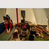 Person reading a book inside a hot tent with a wood stove and cooking equipment.