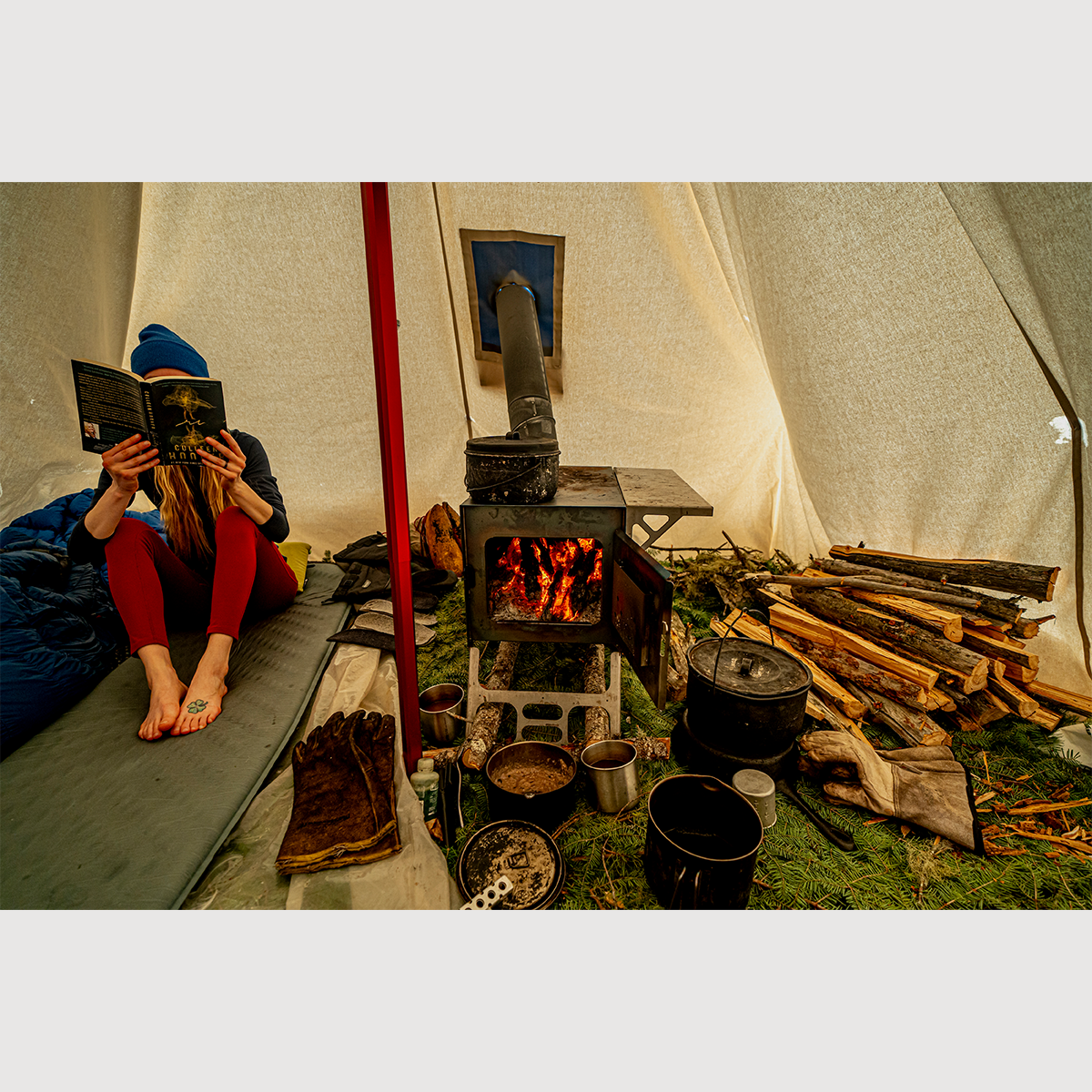 Person reading a book inside a hot tent with a wood stove and cooking equipment.