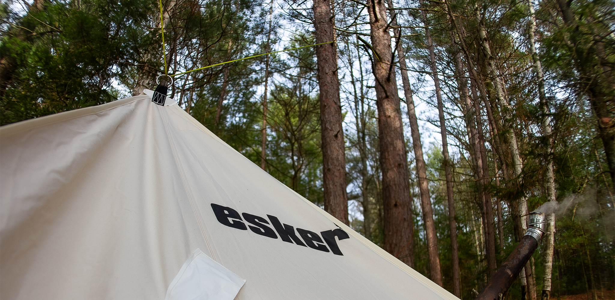 Esker Multi Hot Tent, in the woods with a stove pipe exiting the side wall.